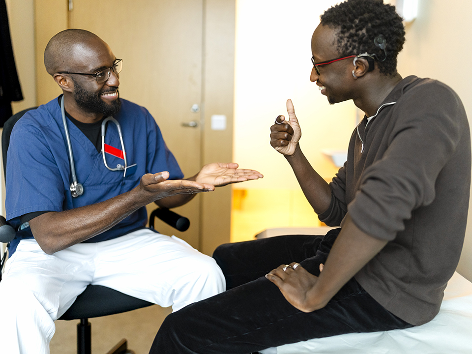 A male doctor seated in a chair engages in conversation with a patient.