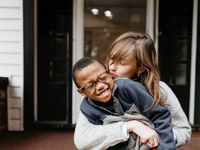 A mother playfully wraps her arms around her son and kisses his cheek as he laughs and leans towards his side
