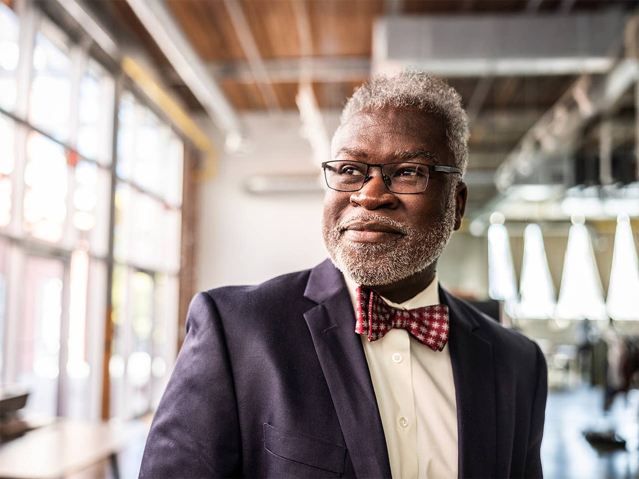 Elderly Black man wearing a navy blue sport coat with a white shirt and bowtie looks out a window
