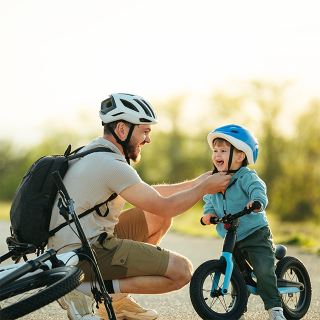 Smiling dad wearing a bike helmet clips a bike helmet on his smiling son while they are outdoors in the sunshine