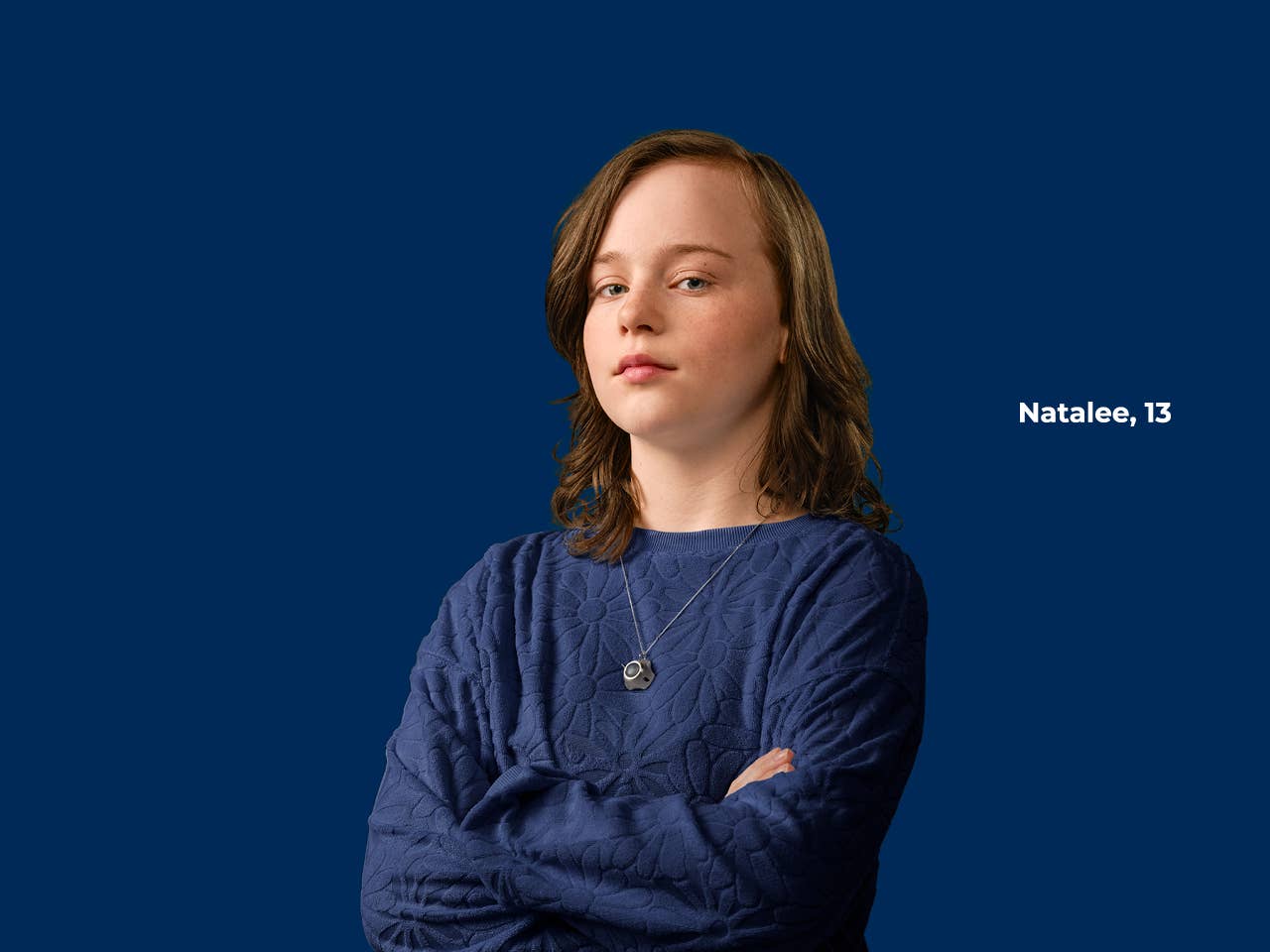 Preteen girl in a dark blue sweater standing with arms folded in front of dark blue background