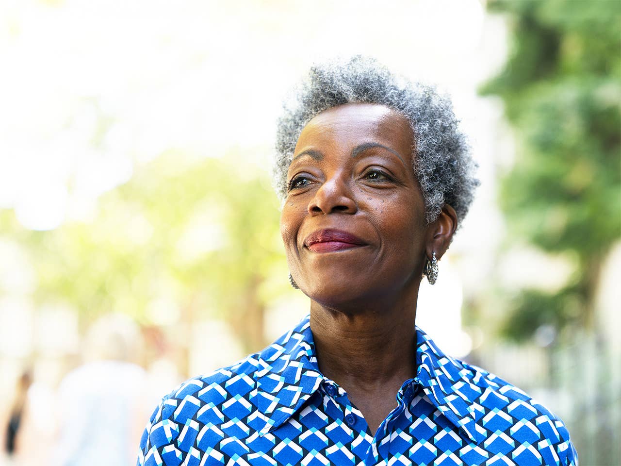 Elderly Black woman wearing a blue shirt with white polka dots stands outside in the sunshine