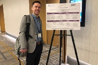 A man stands beside a poster in a hallway, promoting Graduate Medical Education Southeast Michigan Residencies Faculty.