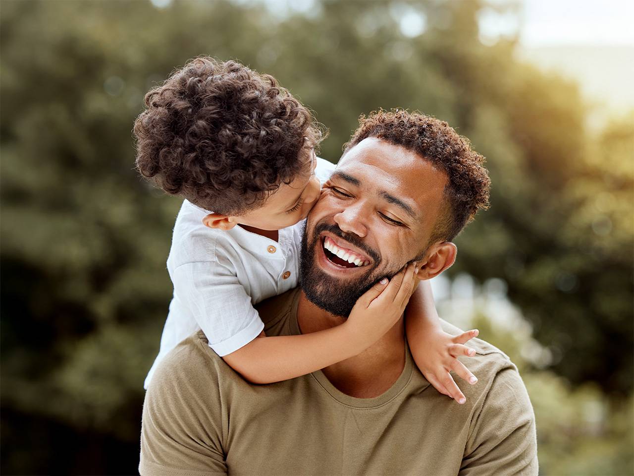 Smiling young Black man carries his son on his back as they play outdoors