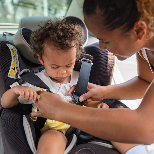 A young mother leans over her toddler while she straps her into a car seat