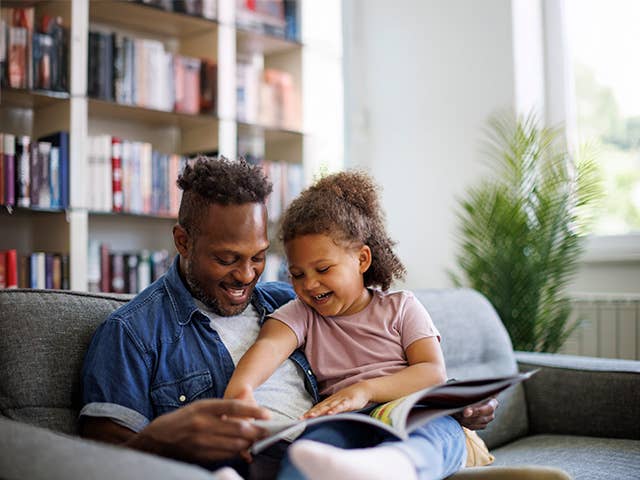 Smiling Black man sits with his young daughter on the sofa, reading a book together