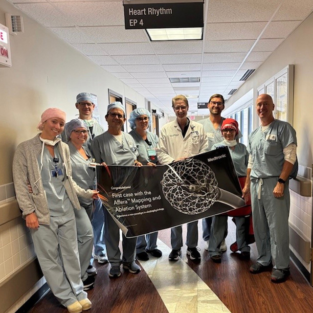 Group of nine Corewell Health heart specialists, a mix of men and women, stand in a hospital hallway holding a banner to celebrate completion of a new atrial fibrillation treatment