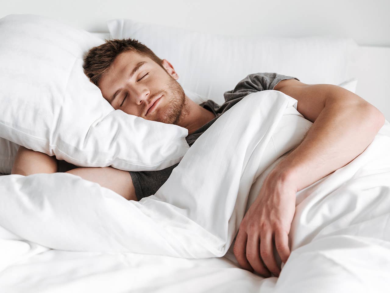 Man with brown hair and a beard sleeps on his side in a bed with white sheets and blankets
