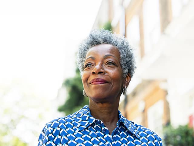 Smiling Black woman with short grey hair stands in the shade outdoors in front of a brick building
