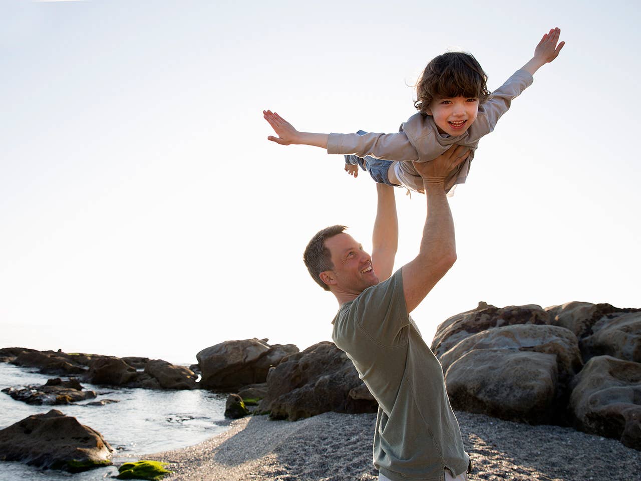 Middle aged man lifts his son into the air alongside the beach
