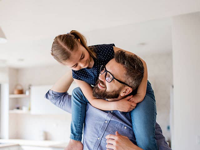 Middle-aged man wearing glasses looks up and smiles at his daughter as she climbs on his back