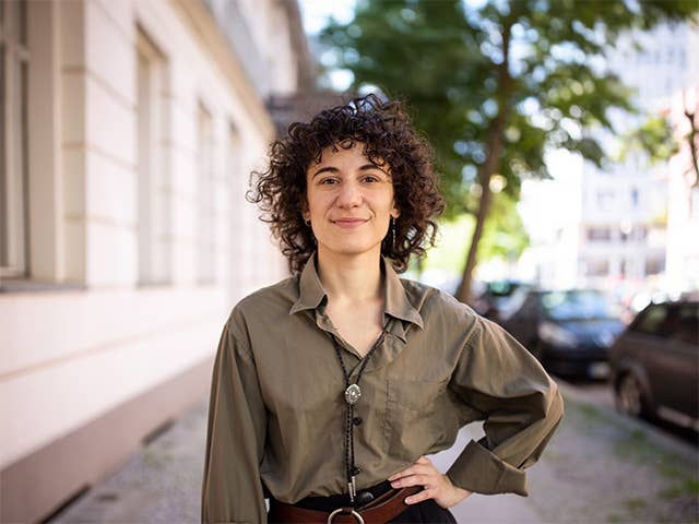 Young white woman with curly brown hair stands on the sidewalk by a building with her hand on her hip