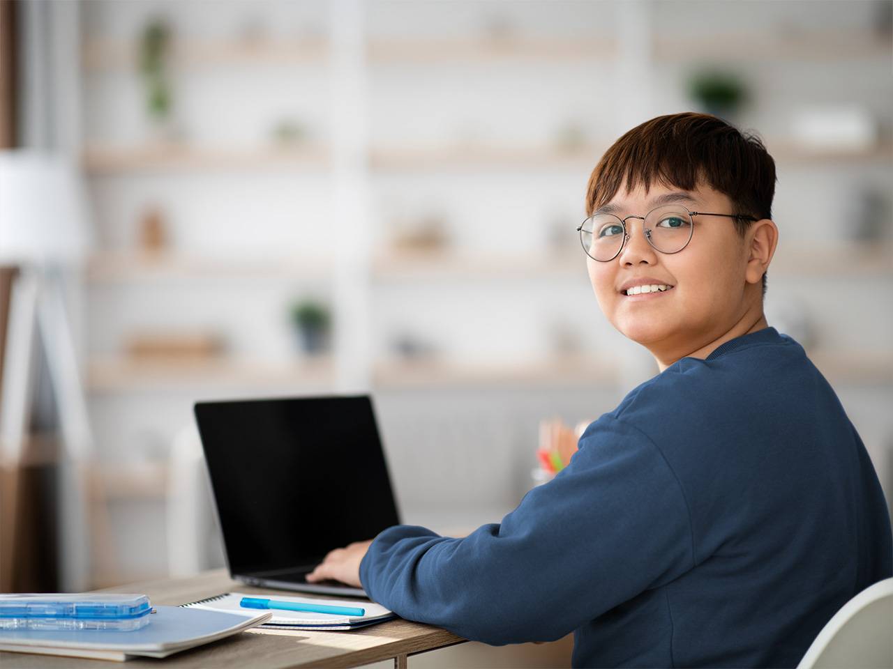A teenage boy with brown hair and glasses turns and smiles while he sits with a laptop at a school desk