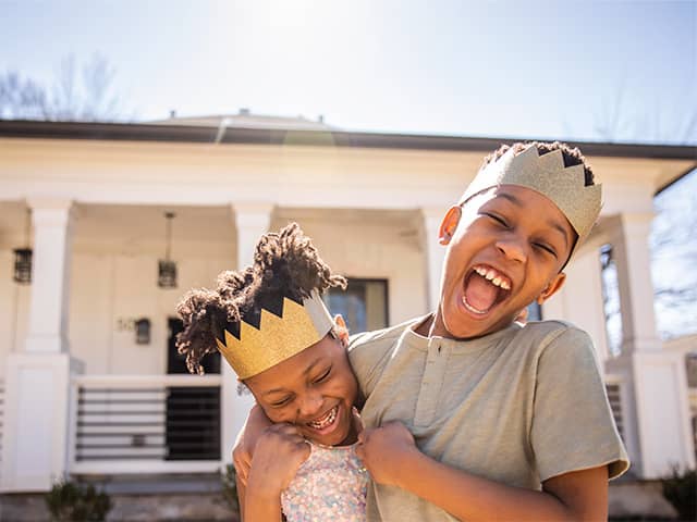 Smiling Black boy wearing a toy crown wraps his arm around his smiling sister as they play outdoors