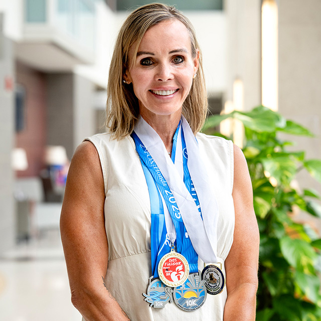 Christine Ankenbauer, a white woman with long blonde hair, wearing a white shirt and five running medals around her neck, standing in front of a hospital building