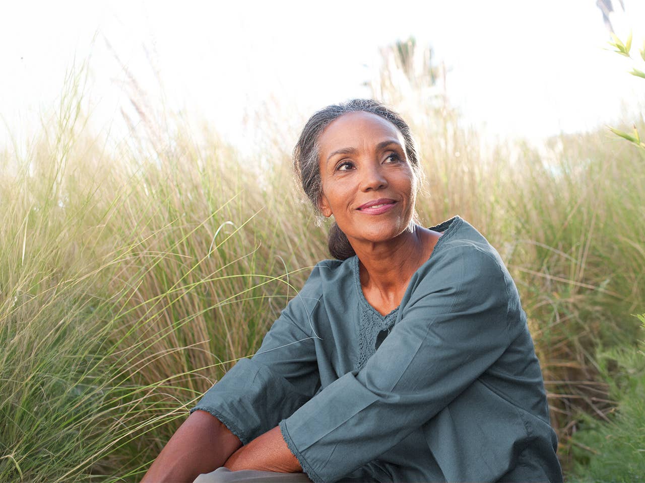 Black woman in a blue shirt sits in long wild grasses in the sunshine