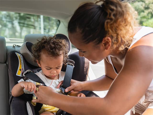 A young mother leans over her toddler while she straps her into a car seat