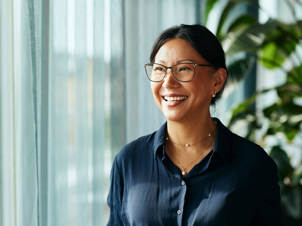 A smiling woman wearing glasses stands in front of a window, with natural light illuminating her face.