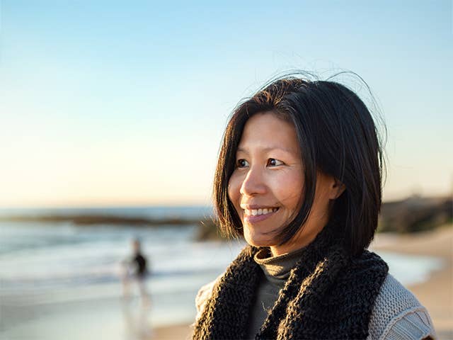 A woman smiles brightly while standing on a sandy beach with gentle waves in the background.