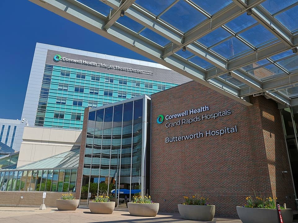 Exterior view of Corewell Health Grand Rapids Hospitals Butterworth Hospital entrance with glass canopy, brick wall signage, and modern multi-story building in the background under a blue sky.
