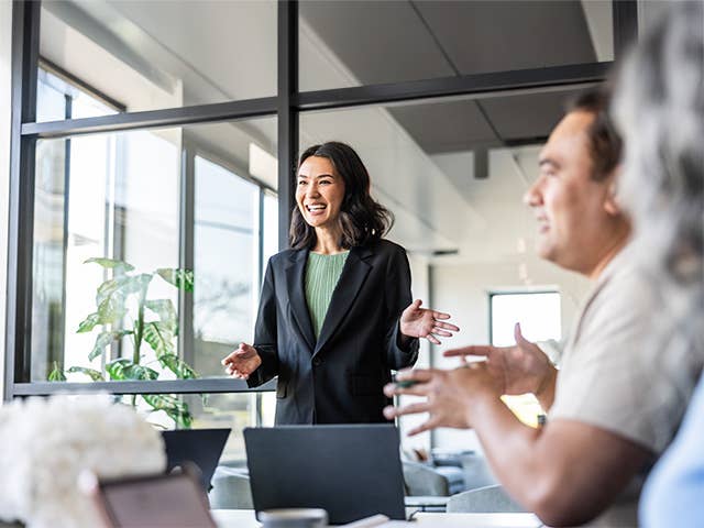 Smiling Asian woman wearing a green blouse and black sport coat stands and leads a discussion in a board room meeting