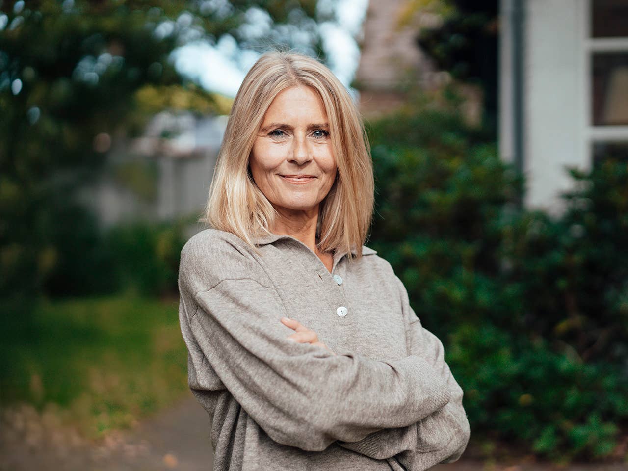 White woman wearing a tan sweater crosses her arms as she stands outside in front of trees