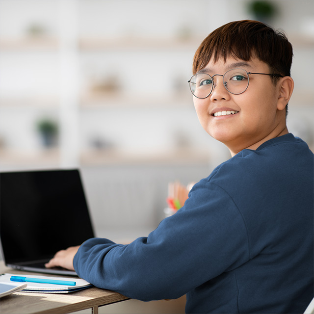 A teenage boy with brown hair and glasses turns and smiles while he sits with a laptop at a school desk