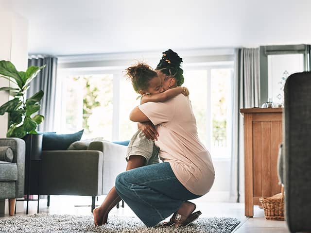 A Black woman kneels down and hugs her young daughter while they are in a sunny living room