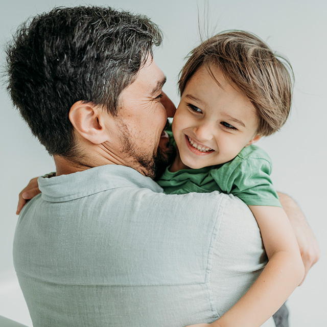 A smiling young boy with brown hair and wearing a green shirt is lifted up by his dad