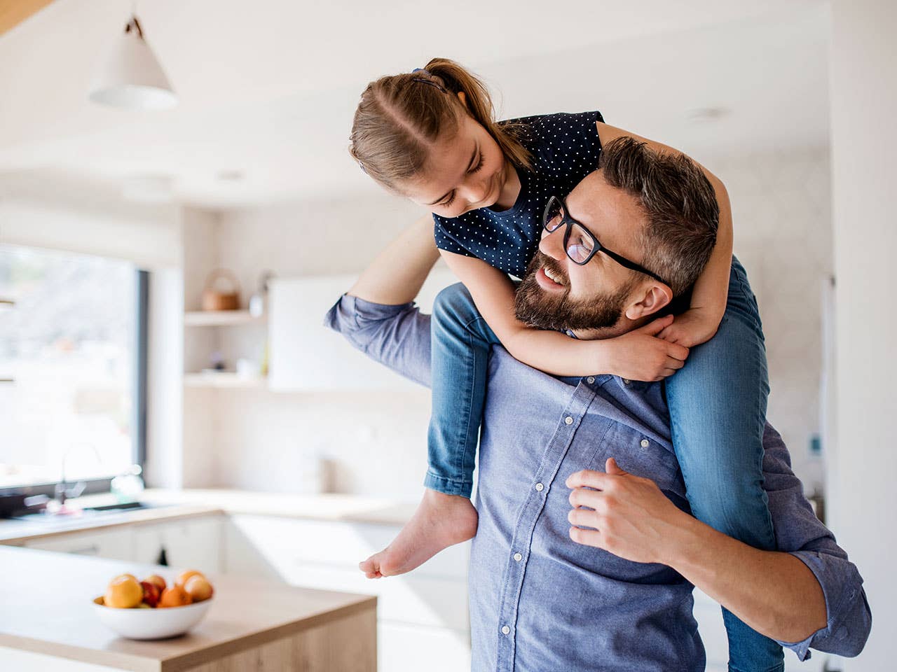 Middle-aged man wearing glasses looks up and smiles at his daughter as she climbs on his back