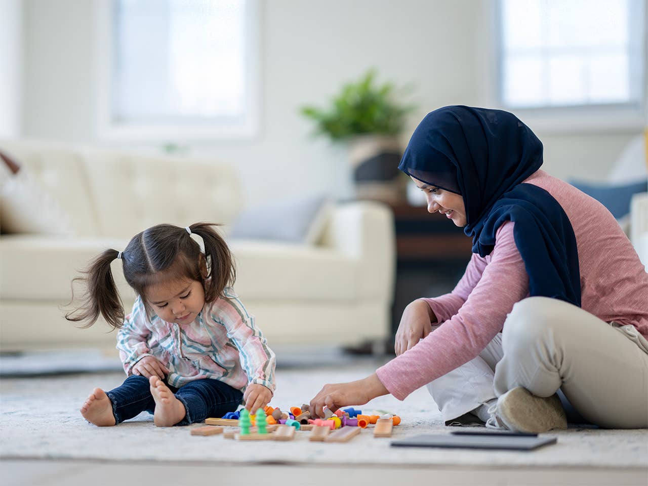 Smiling woman sits on the floor next to her young daughter, playing with toys