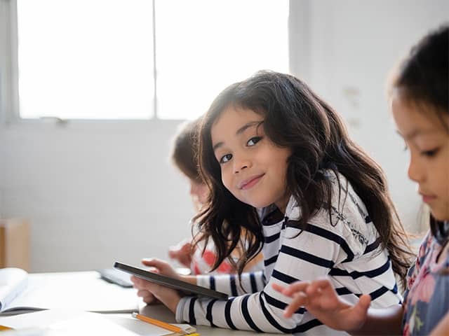 Smiling girl with long brown hair holds a tablet device while in a classroom