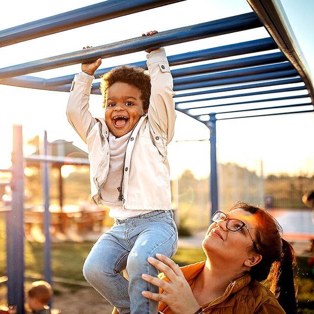 Smiling young Black boy wearing jeans and a white jacket hangs from monkey bars on a playground