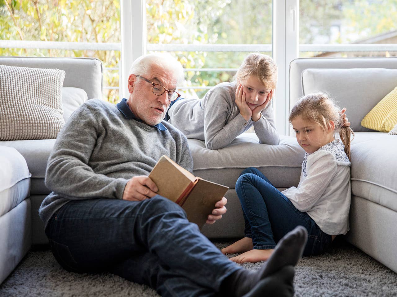 Elderly white man sits on the floor and reads a book to his two granddaughters