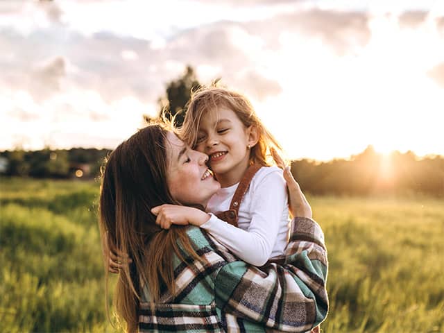 Smiling woman wearing a plaid shirt lifts and hugs her smiling daughter outdoors in the sunshine