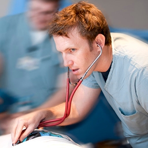 A man in a hospital gown sits on a bed, looking thoughtful in a medical environment, surrounded by clinical equipment.