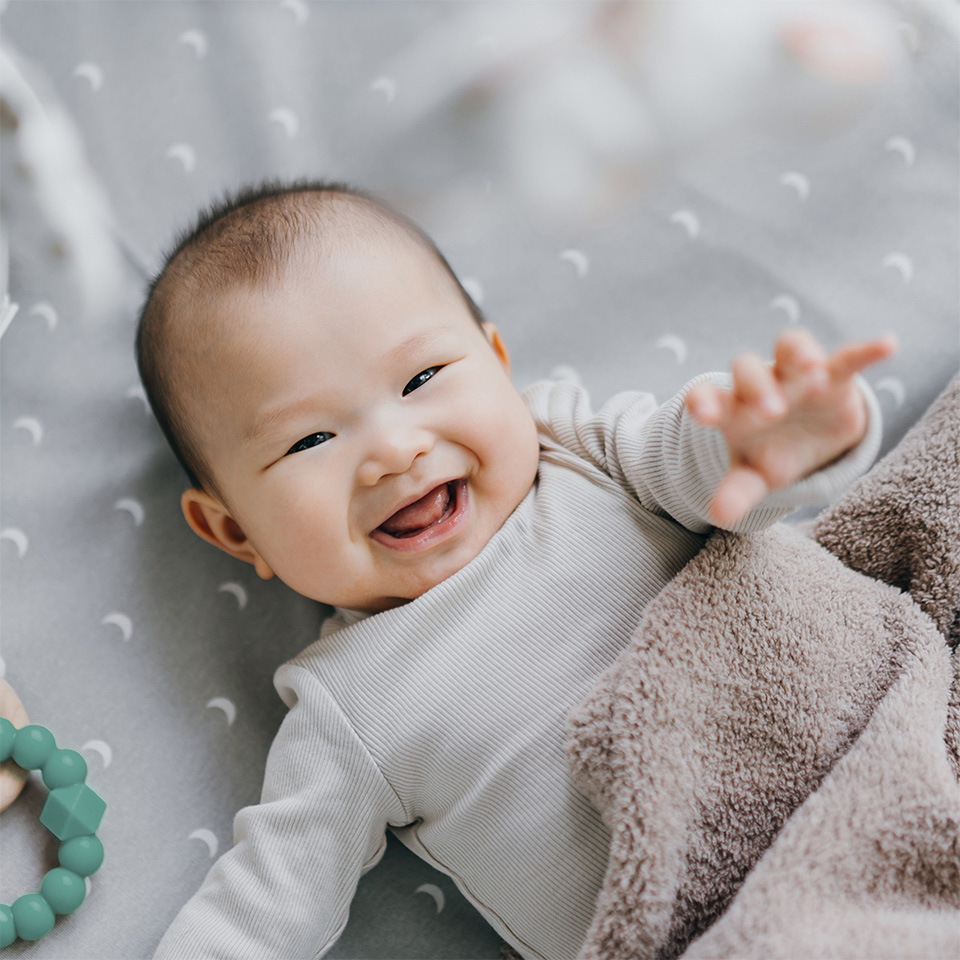 Smiling infant lying on a blanket, reaching upward while resting in a crib or play area.