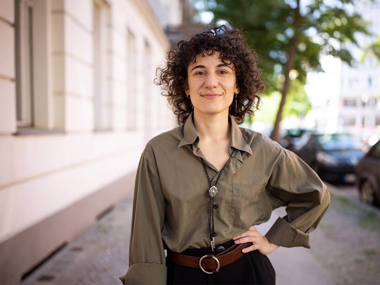 Young white woman with curly brown hair stands on the sidewalk by a building with her hand on her hip
