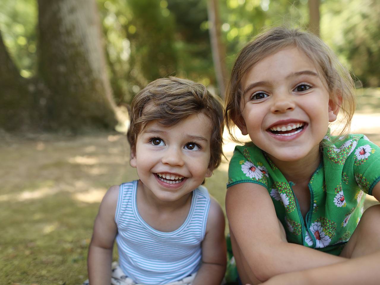Two children sitting close together outdoors in a wooded park setting, facing the camera.