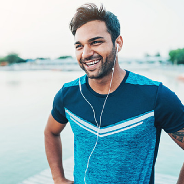 Caucasian man with brown hair, earbuds in ears, wearing a blue shirt, smiles while standing by water