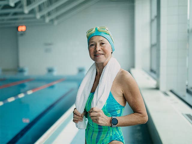 Older woman in a bathing suit and swim cap stands next to a pool with a towel around her neck
