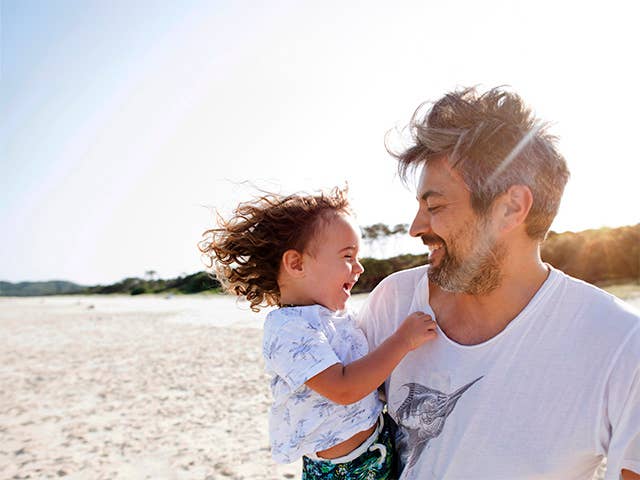 Middle-aged white man holds his toddler son while standing in the sunshine on the beach