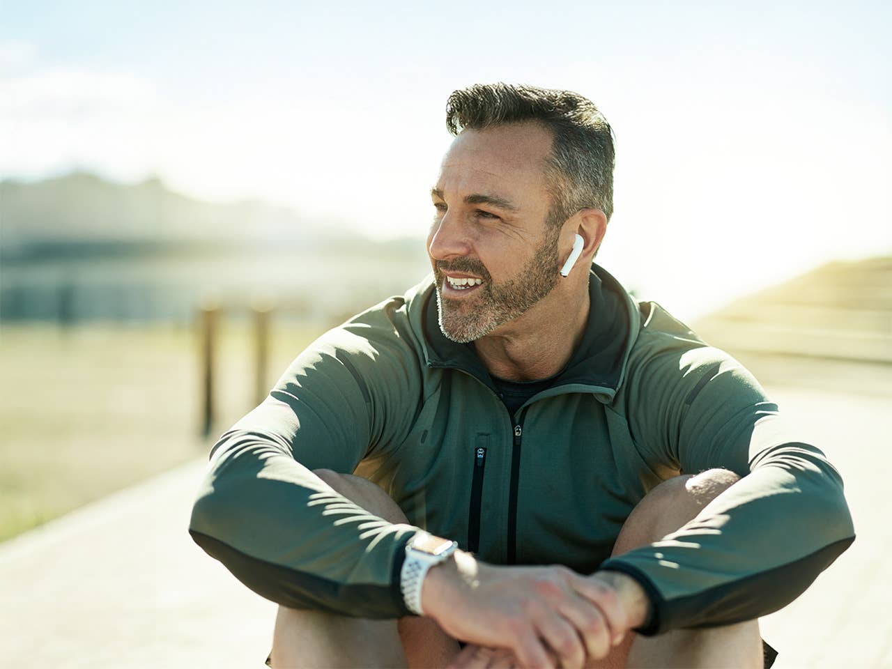 Man wearing a dark green jacket sits in the sunshine wearing earbuds