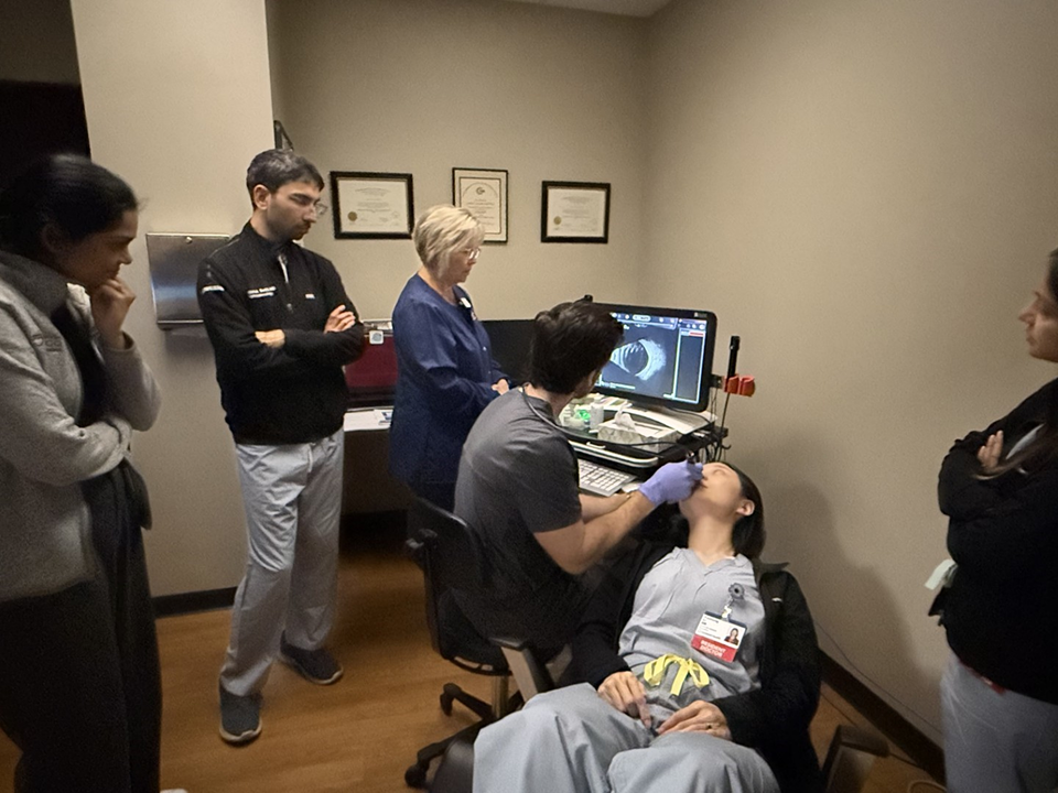 Clinician performs an ultrasound procedure on a patient while several healthcare staff observe the monitor in an exam room.