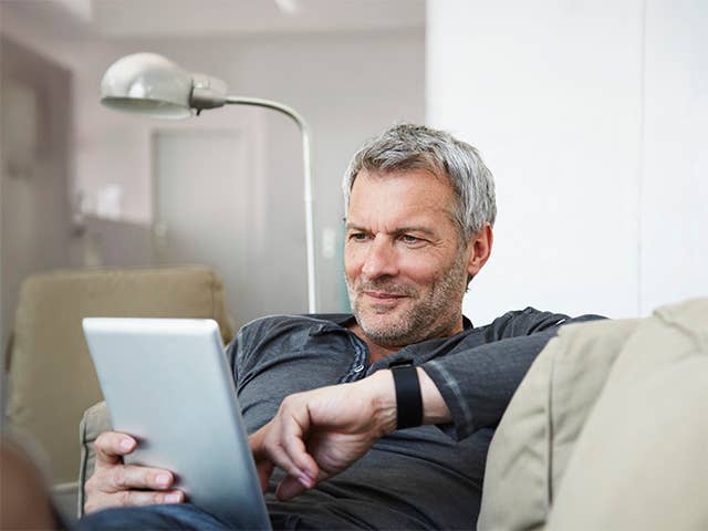 Grey-haired white man sits on a couch looking at a tablet he is holding