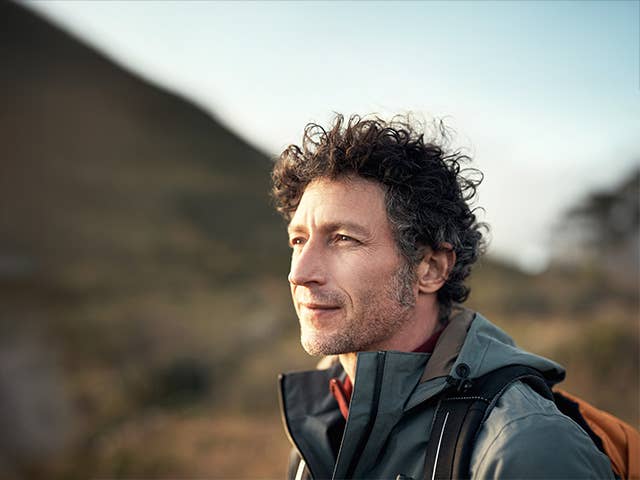 Middle aged white man with curly brown and grey hair stands in the evening sun with a mountain in the background