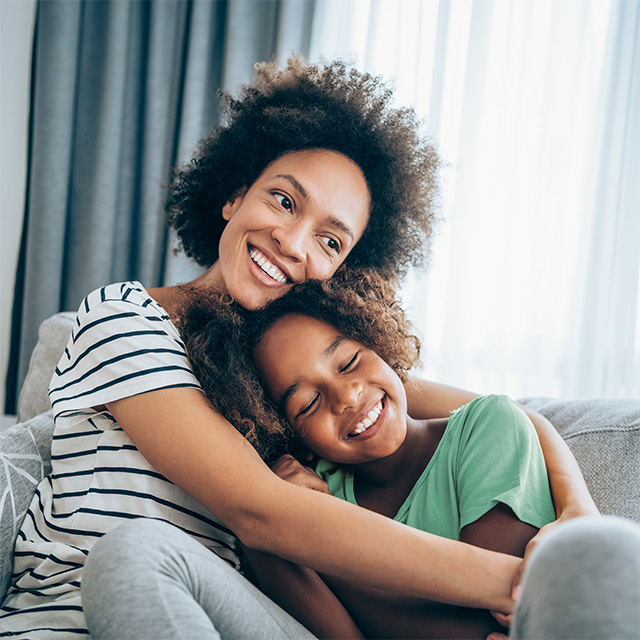 Smiling Black woman wearing a white shirt with navy blue stripes sits on a couch and hugs her smiling daughter