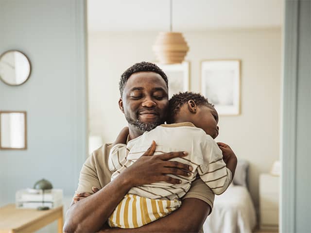 Smiling Black man with dark brown hair and a beard lifts up and hugs his young son