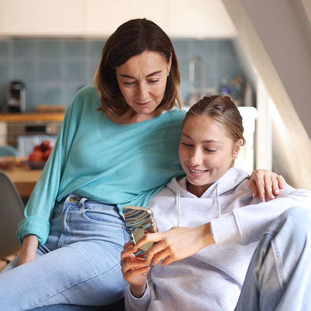 Mother with brown hair and a light blue shirt sits next to her smiling teenage daughter as they look at her phone