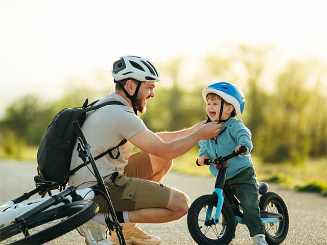 Smiling dad wearing a bike helmet clips a bike helmet on his smiling son while they are outdoors in the sunshine